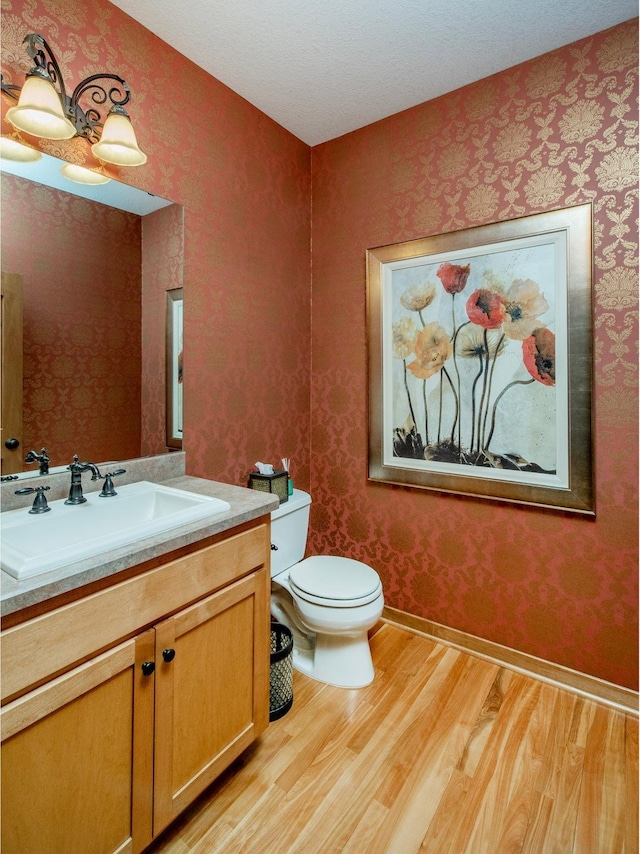 half bathroom with vanity, wood finished floors, toilet, and wallpapered walls