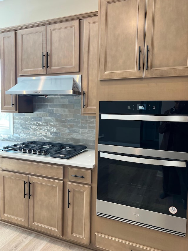 kitchen featuring dobule oven black, decorative backsplash, light countertops, under cabinet range hood, and stainless steel gas stovetop