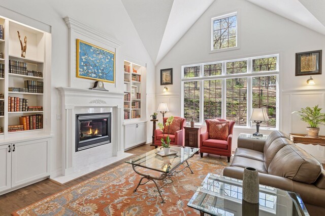 living area featuring built in shelves, a decorative wall, a fireplace with flush hearth, wood finished floors, and wainscoting
