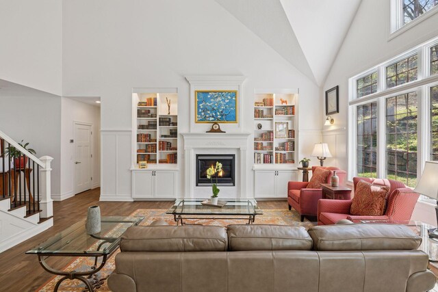 living area with high vaulted ceiling, stairway, wood finished floors, a glass covered fireplace, and built in features
