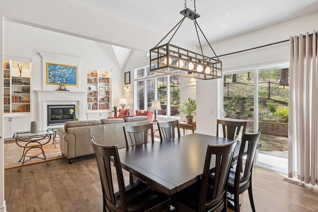 dining area with vaulted ceiling, wood finished floors, a glass covered fireplace, and built in shelves