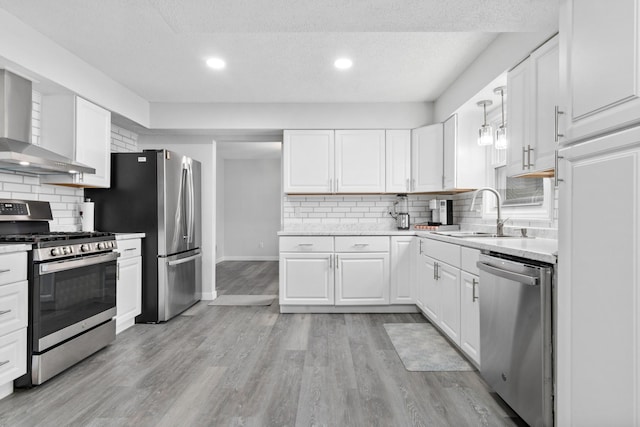 kitchen with light wood finished floors, wall chimney exhaust hood, appliances with stainless steel finishes, white cabinetry, and a sink