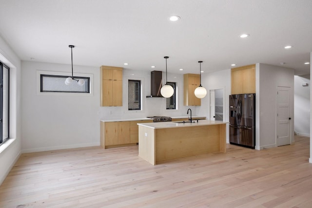 kitchen featuring wall chimney exhaust hood, a kitchen island with sink, black fridge, light brown cabinets, and a sink