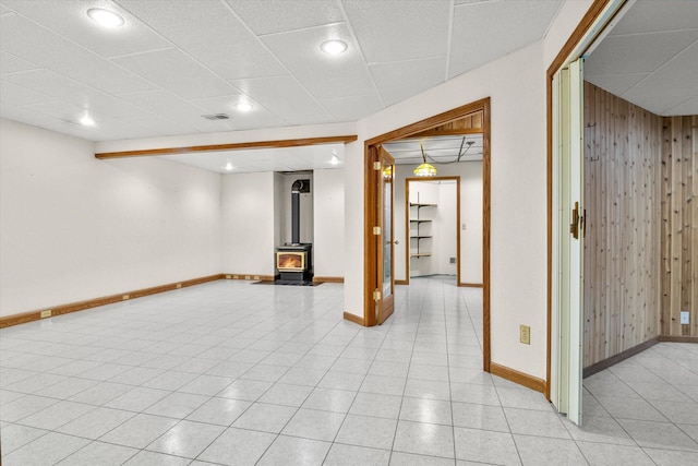 finished below grade area featuring a paneled ceiling, visible vents, a wood stove, wood walls, and baseboards