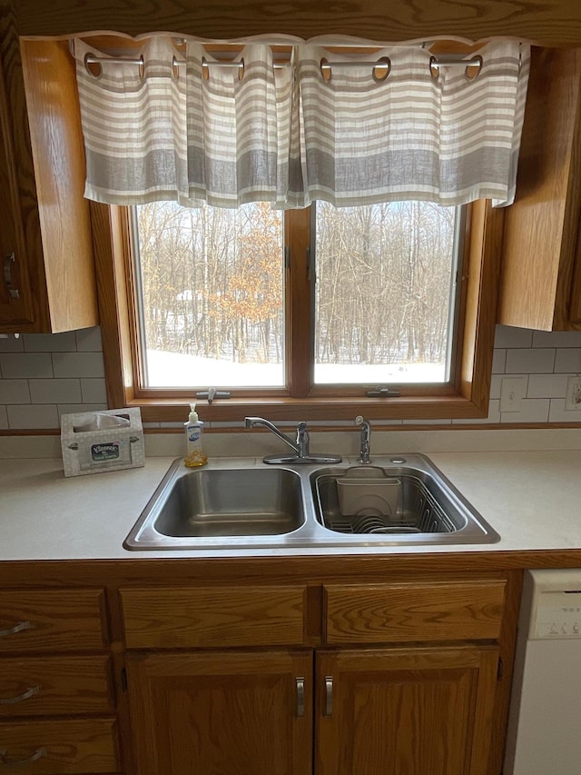 kitchen featuring white dishwasher, a sink, light countertops, decorative backsplash, and brown cabinetry