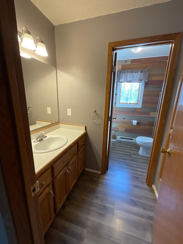 half bathroom with toilet, a baseboard heating unit, vanity, a textured ceiling, and wood finished floors