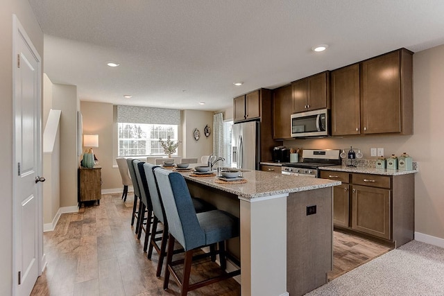 kitchen featuring appliances with stainless steel finishes, light wood-style floors, a kitchen island with sink, light stone countertops, and a kitchen bar