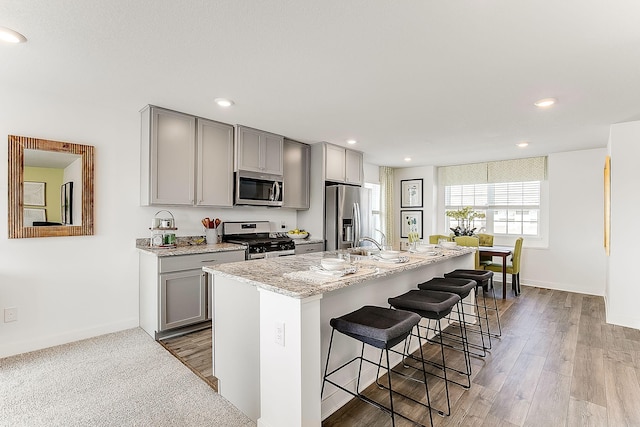 kitchen with a center island with sink, appliances with stainless steel finishes, light stone countertops, gray cabinetry, and a kitchen bar