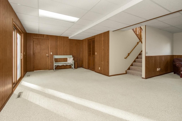 finished below grade area featuring visible vents, carpet flooring, wooden walls, a drop ceiling, and stairs