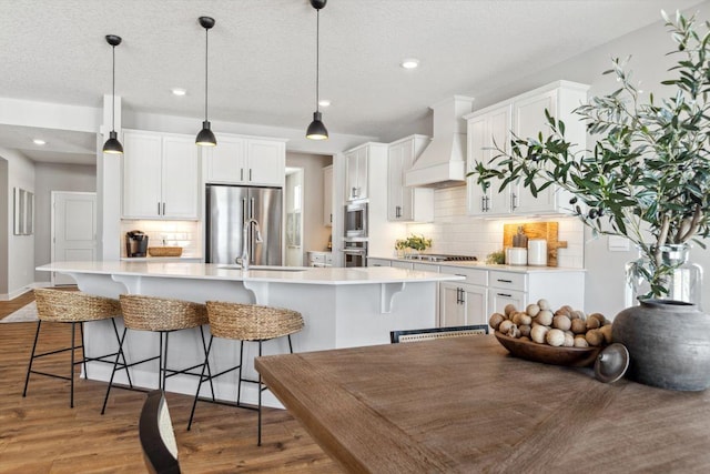 kitchen featuring premium range hood, light wood-style flooring, a kitchen island with sink, light countertops, and appliances with stainless steel finishes