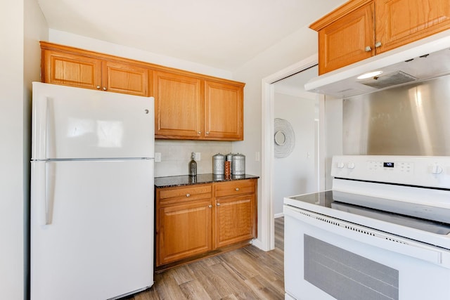 kitchen with brown cabinetry, dark stone counters, light wood-type flooring, white appliances, and under cabinet range hood