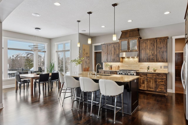 kitchen with hanging light fixtures, glass insert cabinets, a sink, light stone countertops, and under cabinet range hood