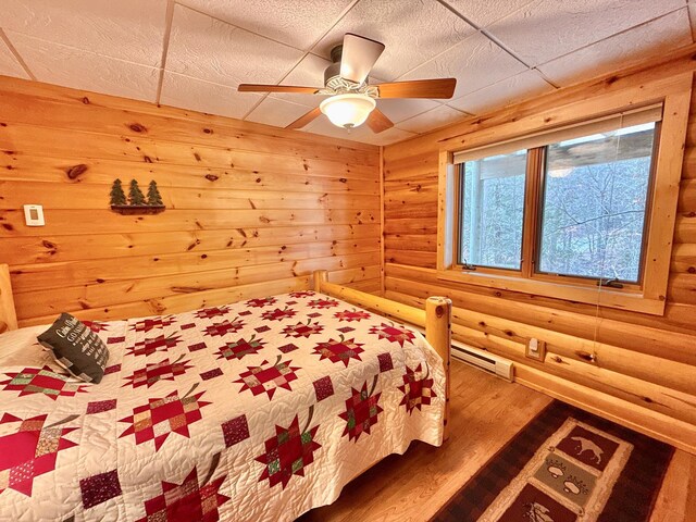 bedroom with log walls, wood finished floors, a baseboard heating unit, and a ceiling fan
