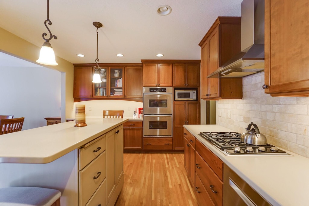 kitchen featuring a breakfast bar area, light countertops, glass insert cabinets, appliances with stainless steel finishes, and wall chimney exhaust hood