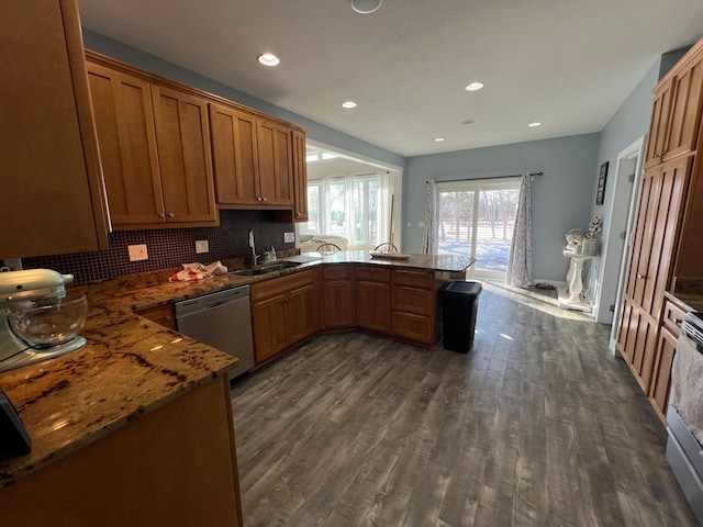 kitchen with a peninsula, a sink, stainless steel dishwasher, dark stone counters, and brown cabinetry