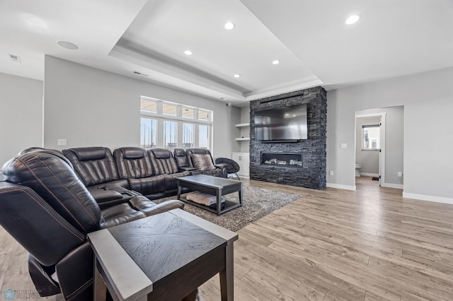 living room featuring a stone fireplace, a tray ceiling, light wood-type flooring, and baseboards