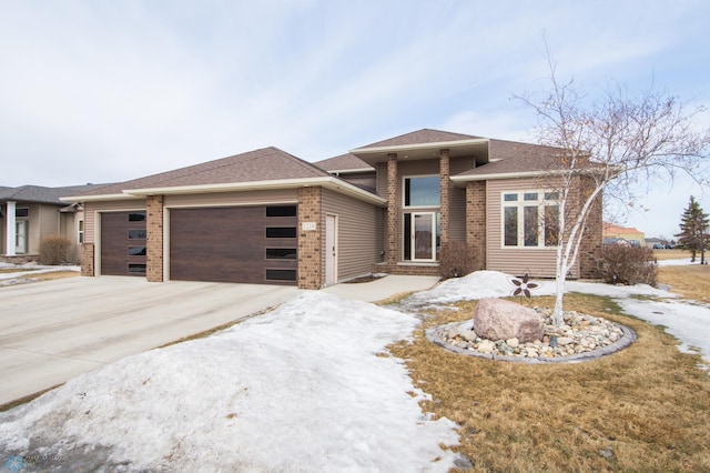 prairie-style house with a garage, driveway, a shingled roof, and brick siding