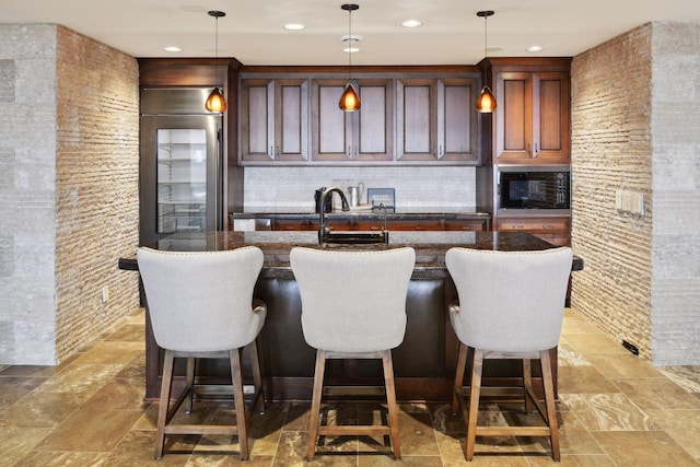 kitchen with a sink, a center island with sink, decorative backsplash, stone finish floor, and decorative light fixtures