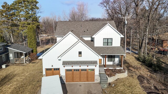 modern inspired farmhouse featuring an attached garage, covered porch, a shingled roof, driveway, and a front lawn