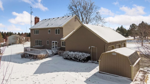 snow covered back of property with an outbuilding, a storage shed, an attached garage, a wooden deck, and a chimney