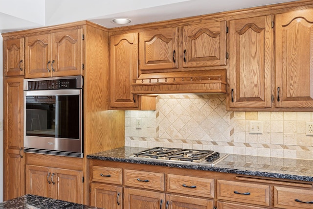 kitchen with dark stone countertops, brown cabinetry, custom exhaust hood, decorative backsplash, and appliances with stainless steel finishes