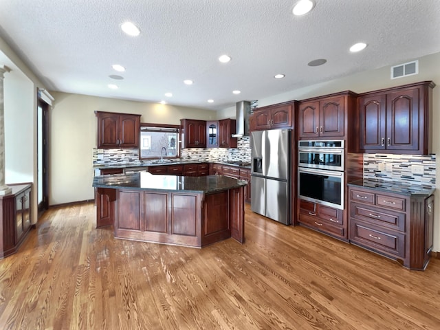 kitchen featuring wall chimney exhaust hood, wood finished floors, dark stone countertops, a center island, and stainless steel appliances