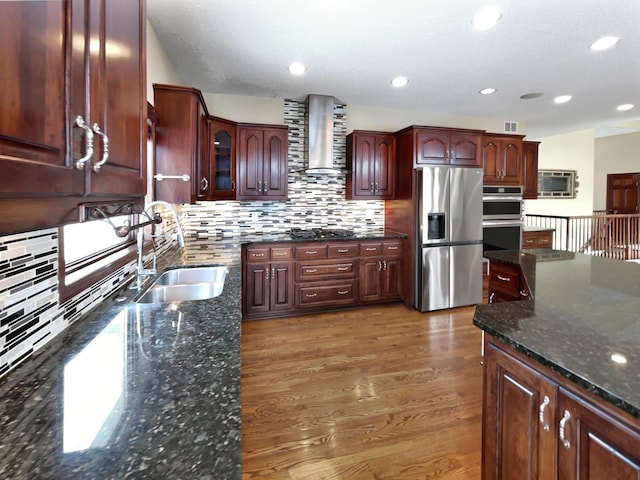 kitchen with dark wood-type flooring, dark stone countertops, stainless steel appliances, wall chimney range hood, and a sink