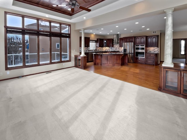 unfurnished living room with light wood-style flooring, visible vents, a ceiling fan, a raised ceiling, and ornate columns