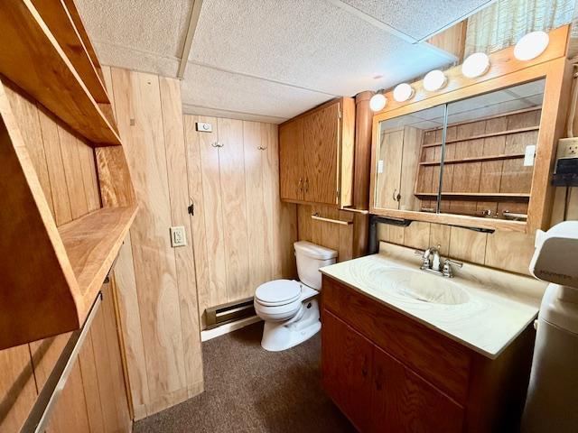 half bathroom with a baseboard radiator, wood walls, vanity, and toilet