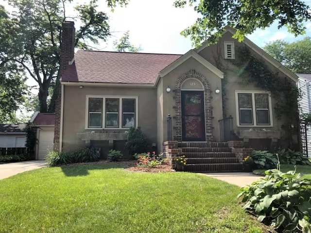 view of front facade featuring a front lawn, concrete driveway, roof with shingles, stucco siding, and a chimney