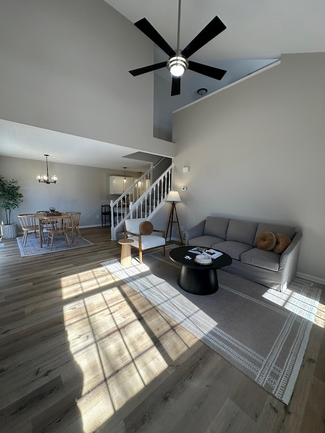 unfurnished living room with high vaulted ceiling, ceiling fan with notable chandelier, wood finished floors, baseboards, and stairs