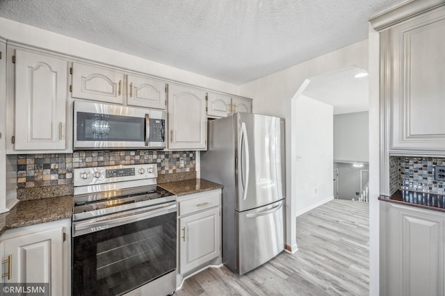 kitchen with light wood finished floors, tasteful backsplash, dark stone counters, appliances with stainless steel finishes, and white cabinetry