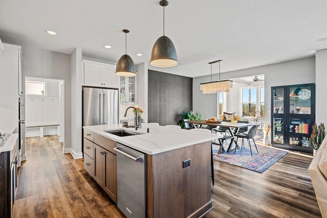 kitchen with a kitchen island with sink, stainless steel appliances, a sink, hanging light fixtures, and dark wood finished floors