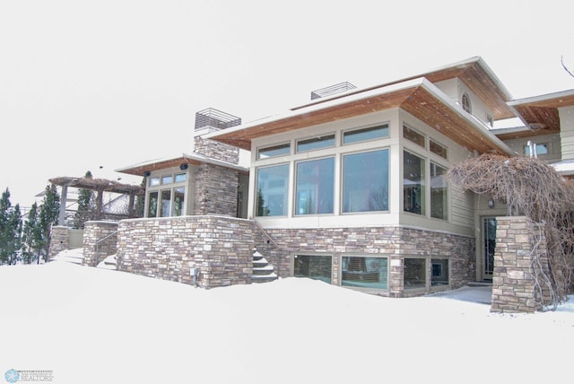 view of snow covered exterior featuring stairs, stone siding, a chimney, and a sunroom