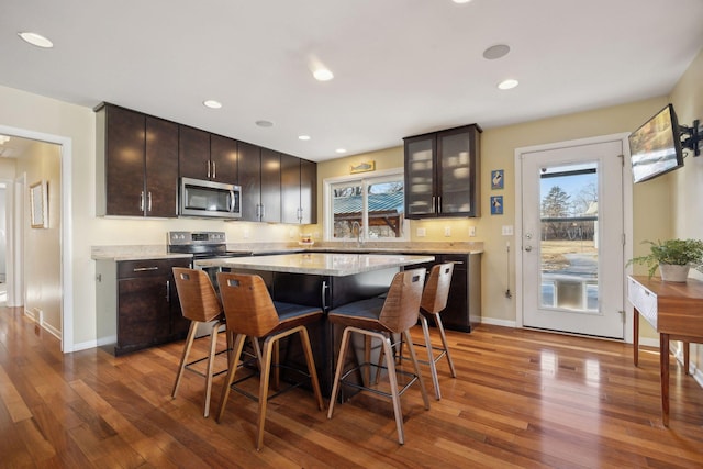 kitchen with a breakfast bar area, stainless steel appliances, glass insert cabinets, a kitchen island, and wood finished floors