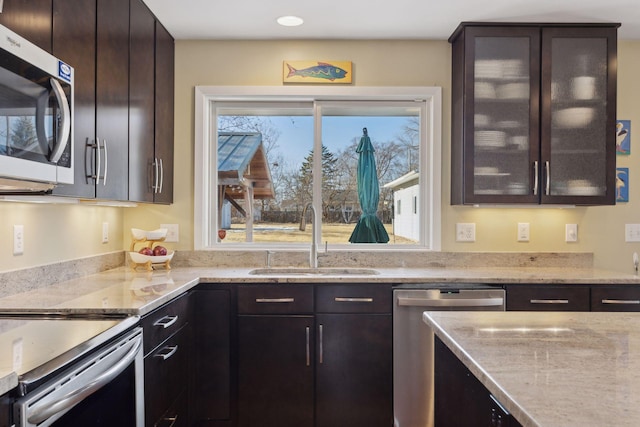 kitchen with glass insert cabinets, light stone countertops, stainless steel appliances, dark brown cabinets, and a sink