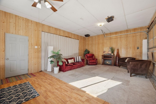 living room featuring a paneled ceiling, a ceiling fan, wood walls, wood finished floors, and a garage