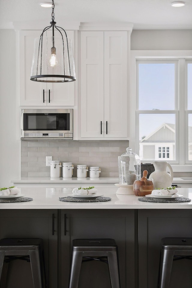 kitchen featuring stainless steel microwave, decorative light fixtures, light countertops, white cabinetry, and backsplash