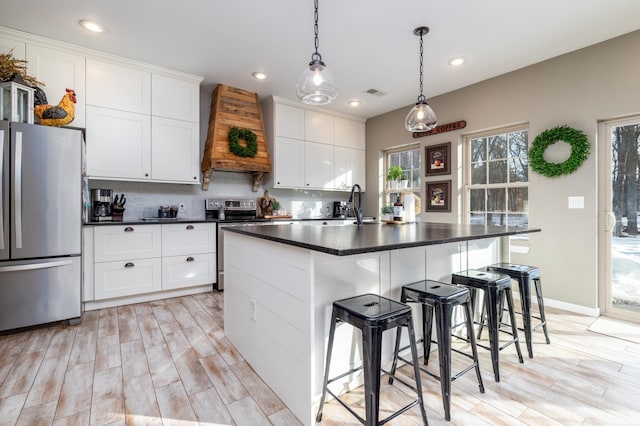 kitchen with a center island with sink, dark countertops, custom range hood, appliances with stainless steel finishes, and white cabinets