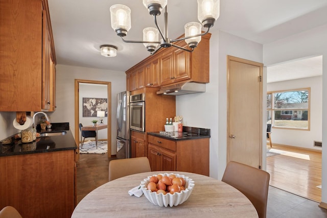 kitchen featuring brown cabinetry, stainless steel appliances, a sink, under cabinet range hood, and dark countertops