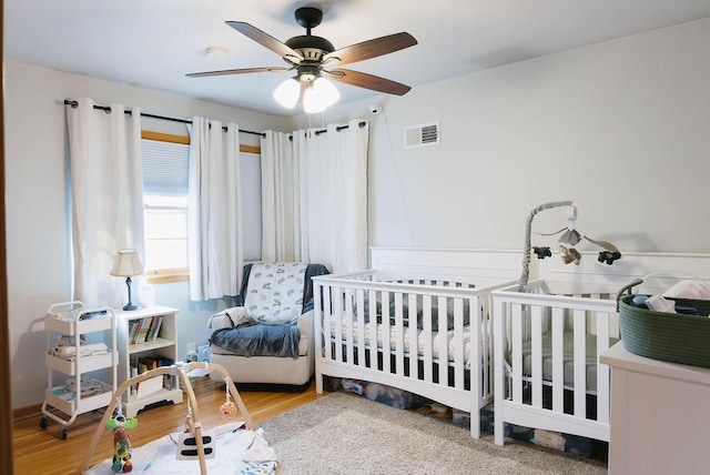 bedroom with ceiling fan, a nursery area, wood finished floors, and visible vents