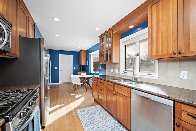 kitchen with brown cabinetry, glass insert cabinets, appliances with stainless steel finishes, dark stone countertops, and a sink