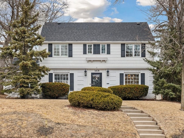 colonial house with brick siding, a chimney, and a shingled roof