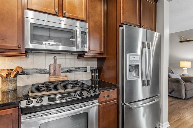 kitchen with stainless steel appliances, wood finished floors, backsplash, dark stone counters, and brown cabinetry