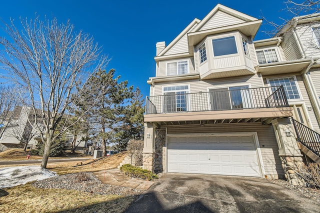 multi unit property with a garage, a balcony, stone siding, a chimney, and driveway