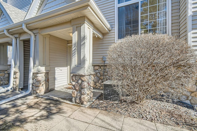 entrance to property featuring a shingled roof, central air condition unit, and stone siding
