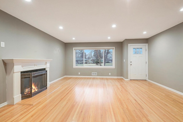 unfurnished living room with a warm lit fireplace, light wood finished floors, recessed lighting, baseboards, and visible vents