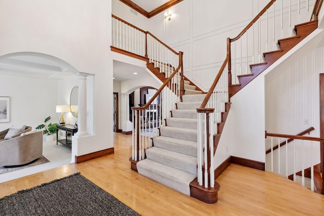 stairway with arched walkways, wood finished floors, a towering ceiling, decorative columns, and crown molding