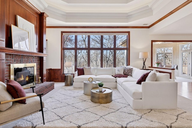 living room with a wainscoted wall, a raised ceiling, ornamental molding, a brick fireplace, and light carpet