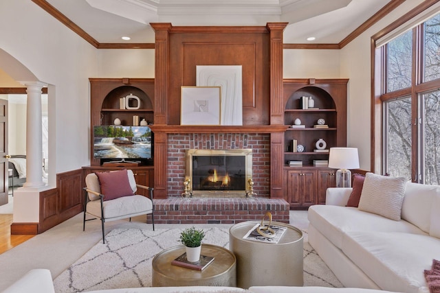 living area featuring crown molding, a brick fireplace, a wainscoted wall, and decorative columns